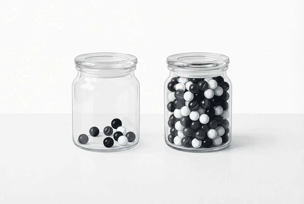 Two glass jars of dark and white marbles, one with a few marbles and one packed full, side by side on a table.